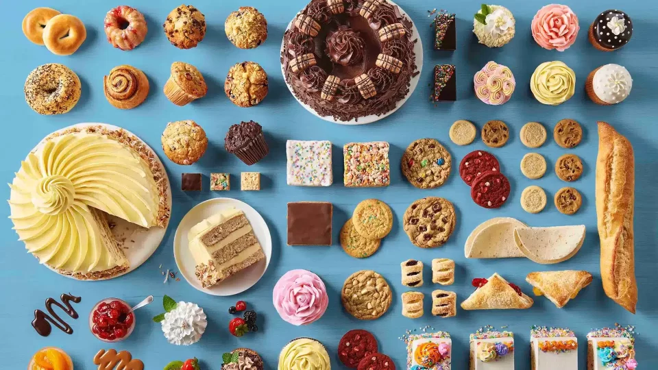 A large variety of baked goods laid out neatly on a table. Muffins, cakes, donuts, bagels, cookies, cupcakes, bread, and pastries are all present.