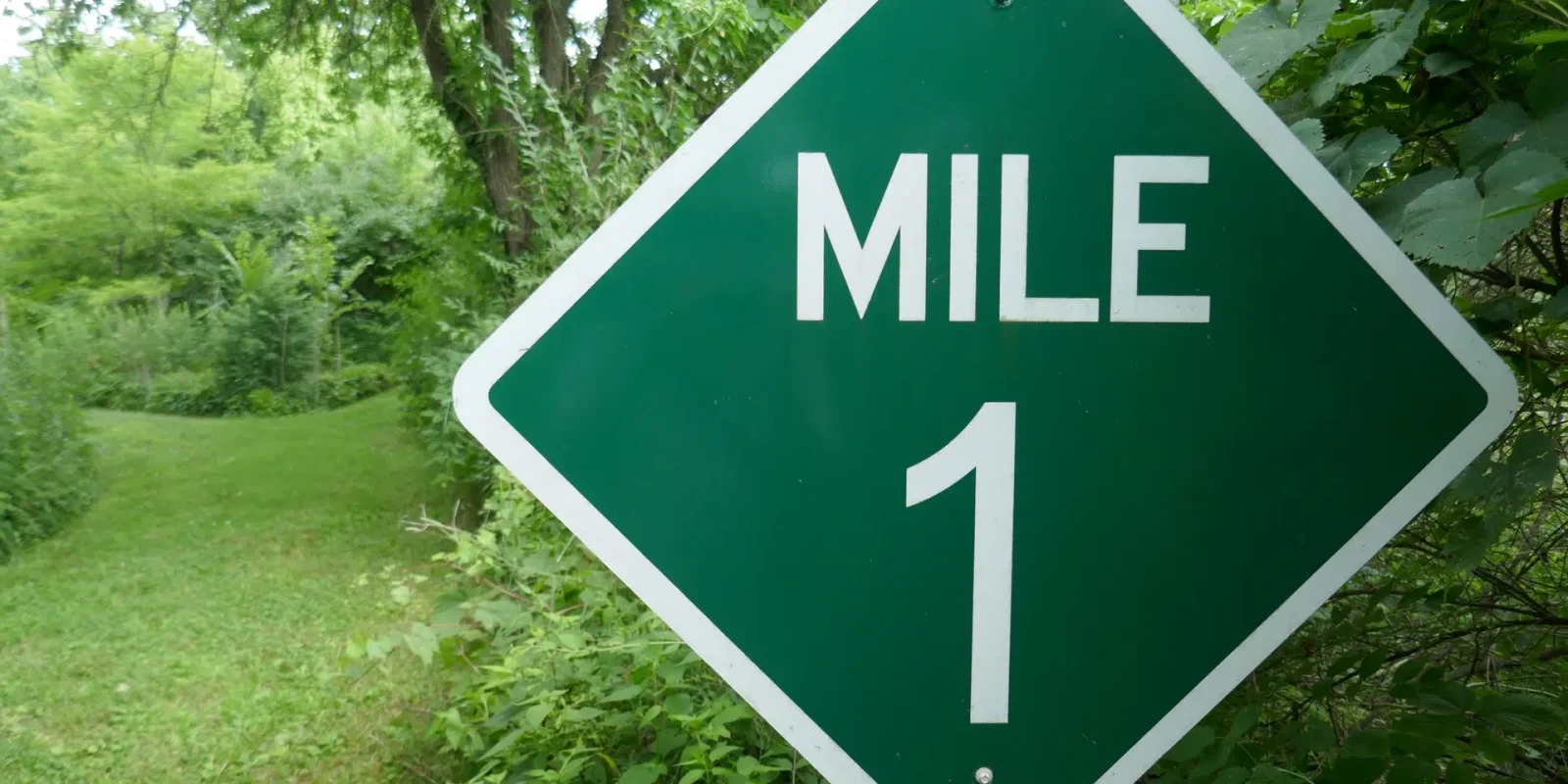 A road sign reading "Mile 1" with trees and greenery in the background.