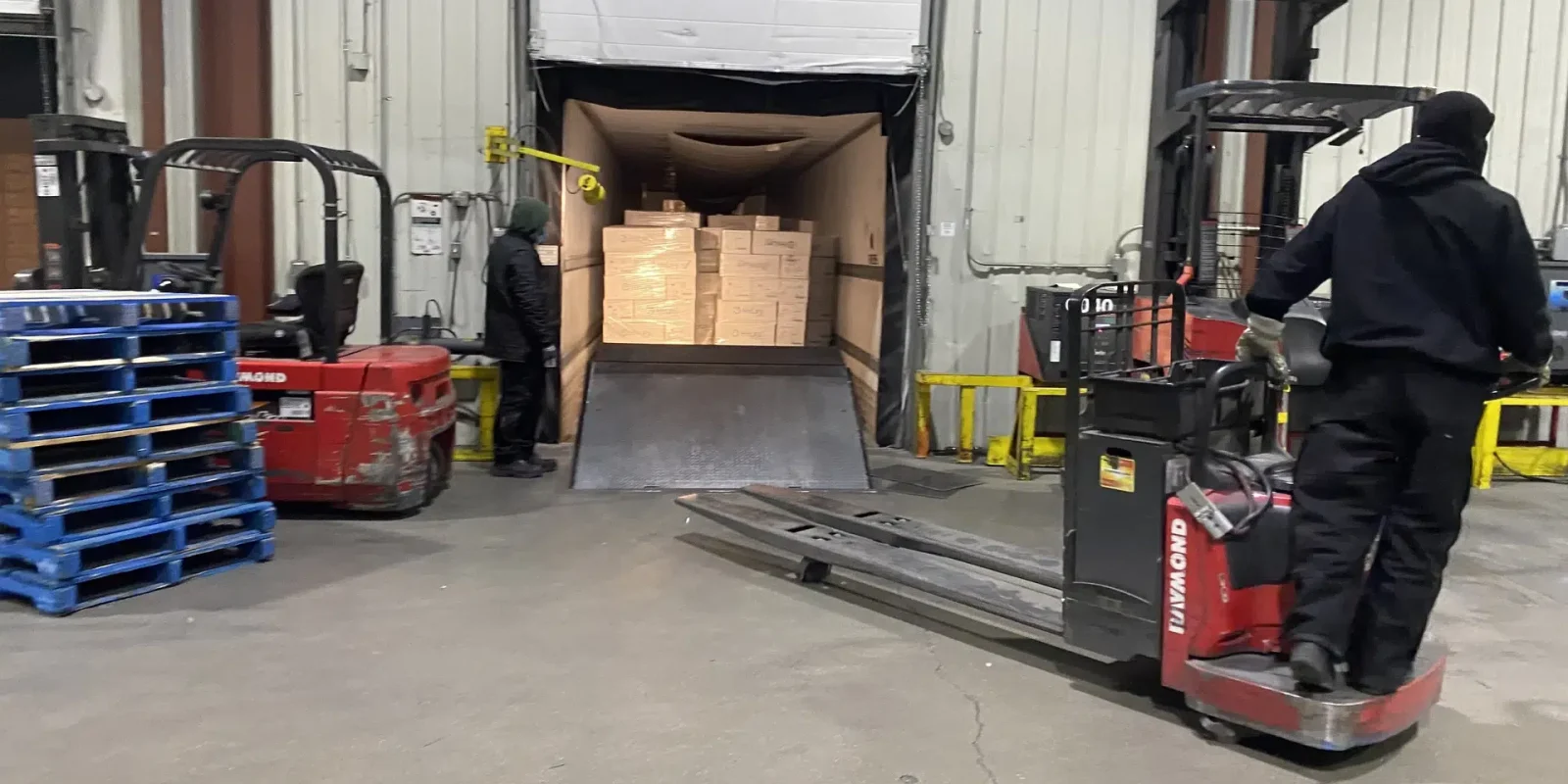 transloading-hylife-manitoba-facility Two Trappers Transport employees transloading freight at the Hylife Manitoba facility. One stands next to an open temperature controlled transport container while the other operates a fork lift.